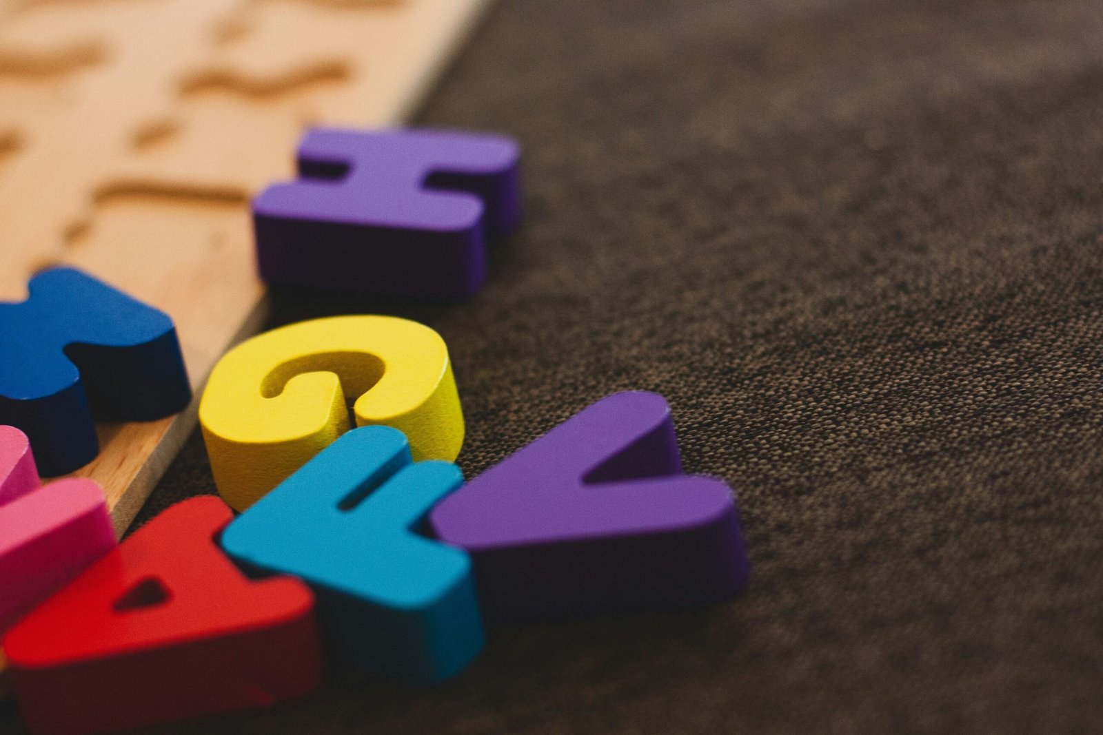Close-up of colorful foam alphabet puzzle pieces on a light wooden tray, scattered on a dark fabric surface.