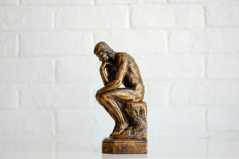 Bronze sculpture of a seated thinker with elbows on knees against a white brick wall backdrop. Non-zoomed shot shows weathered patina on the statue's surface.